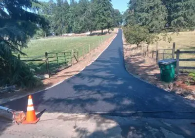 Freshly paved rural driveway flanked by fences, trees, and a green trash bin