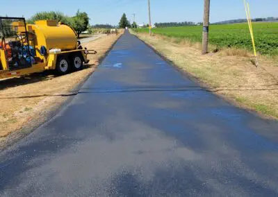 Newly sealed rural road stretching between fields with yellow paving equipment nearby